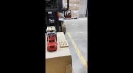 Using a forklift, I place a miniature toy forklift on the forks, pick up a tiny toy pallet, and carefully place it onto another miniature pallet.