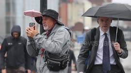 A man uses a miniature pink umbrella to shelter his phone as commuters cross London Bridge during heavy rain in Central London