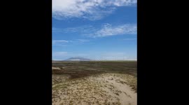 This is a panoramic video of a vast coastal dune or desert landscape under a blue sky with scattered clouds. The camera performs a slow, steady pan from left to right, revealing the immensity of the rolling terrain, which is covered in sand and low, spars