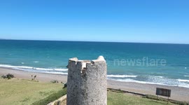 drone flying over old castle style tower turret next to blue sea and clear sky