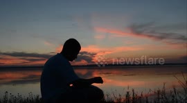 Guy sits on the ground near the river and plays the happy drum. Sunset and calm river
