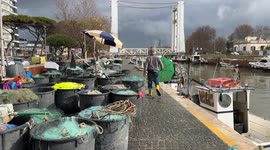 A fisherman in yellow boots walks toward plastic containers holding fishing nets on the fishing boat docks along the canal port on a cloudy December day in Fiumicino.