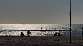 Silhouettes of people on the beach of Lido di Ostia looking at the sea reflecting the sun's rays as a sailboat crosses the scene on the horizon on a sunny Sunday in December in Rome.