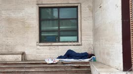 A homeless man sleeps in the warmth of his bed under a canopy sheltered from the wind and rain, while the building opposite is reflected in the glass above, late on a Sunday morning in December in Rome.