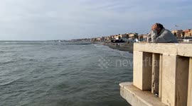 A thoughtful man with his hands on his head leaning with his elbows on the balustrade of the Lido di Ostia pier looks down towards the sea on a sunny October day in Rome.