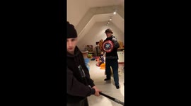 Boys roughhousing around as one swings at a captain America shield, shattering it in the process