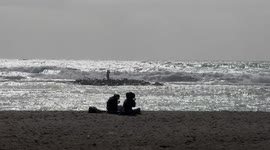 Two people are eating on the beach while in the background there is a very rough sea with reflections of the sun and a cloudy sky on the horizon and a seagull crosses the scene on the coast of Lido di Ostia in Rome.