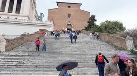 An umbrella vendor looks for customers in front of the Ara Coeli Staircase on a cloudy day in Rome.