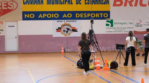 Three young athletes reach the finish line during a children’s indoor inline speed skating competition. Officials and timekeepers observe closely with cameras and timing equipment