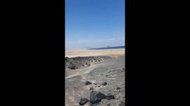 This video, shot from a first-person (POV) perspective, takes the viewer on a hike along an arid, rocky trail overlooking a vast sand plain and a distant body of water. A hand occasionally enters the frame, pointing out the path and scenery. The audio fea