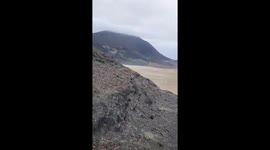This video presents a sweeping panoramic view of an arid coastal landscape under an overcast sky. The camera pans slowly, starting from a rocky hillside, moving across a vast sand plain, and finally revealing a calm bay or sea dotted with small boats. The
