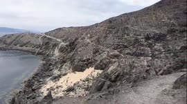 This is a panoramic, high-angle video showing a rocky and arid coastline under an overcast sky. The camera slowly pans from left to right, tracing a narrow path carved into the cliffside before opening up to reveal a vast expanse of sea dotted with numero