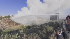 A burning hill of junk made of car parts, metal and wood. A huge water cannon shoots at the fire at 100 liters per second